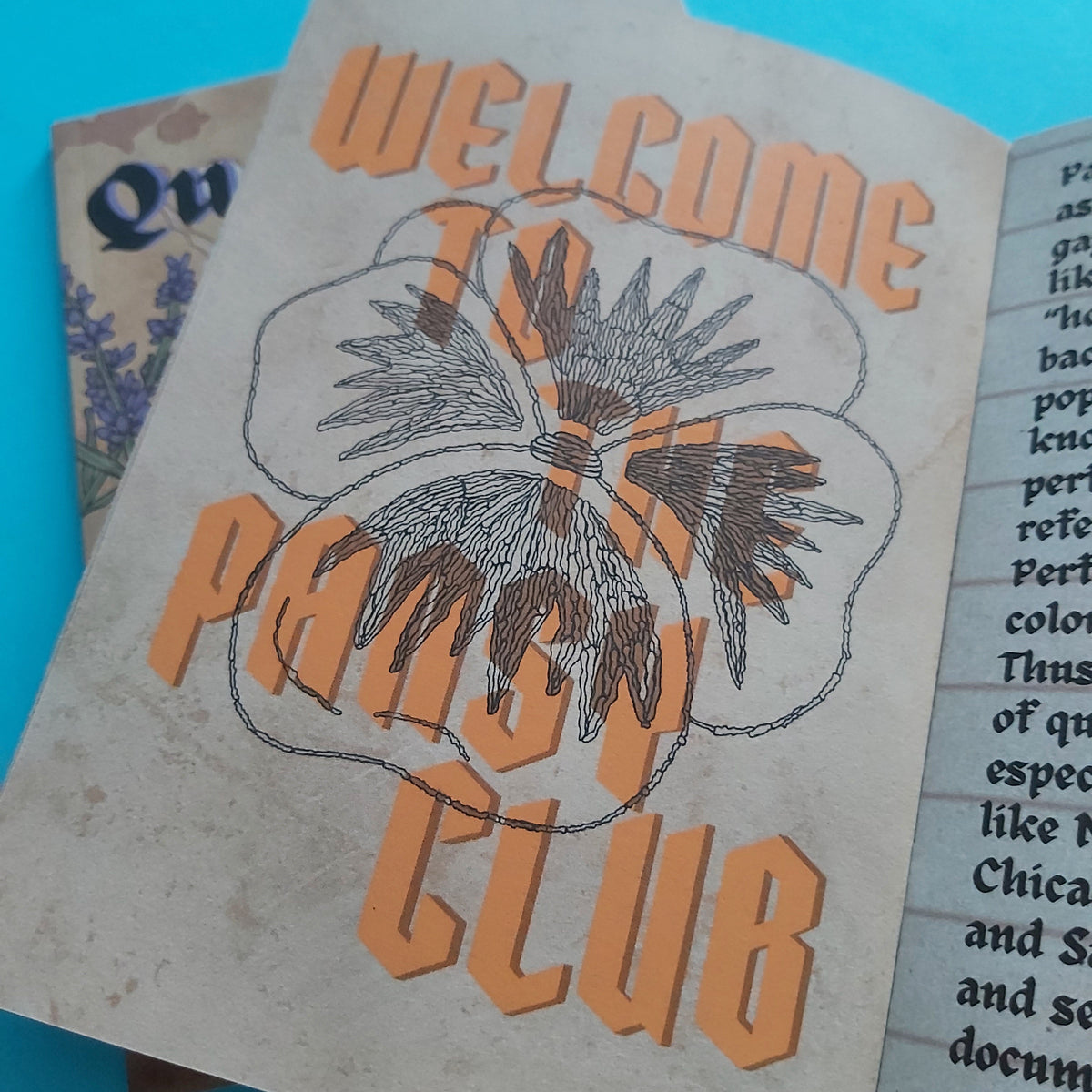 A close-up of a vintage-style printed zine page reading 'WELCOME TO PANSY CLUB' in orange lettering over a black-and-white floral illustration, placed on a bright blue surface with part of another printed page visible underneath.