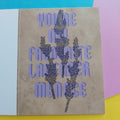 A greeting card with a textured beige background, featuring a purple and black lavender illustration and the text 'YOU'RE MY FAVORITE LAVENDER MENACE' on the front, displayed on a colourful background of yellow, pink, and blue paper.