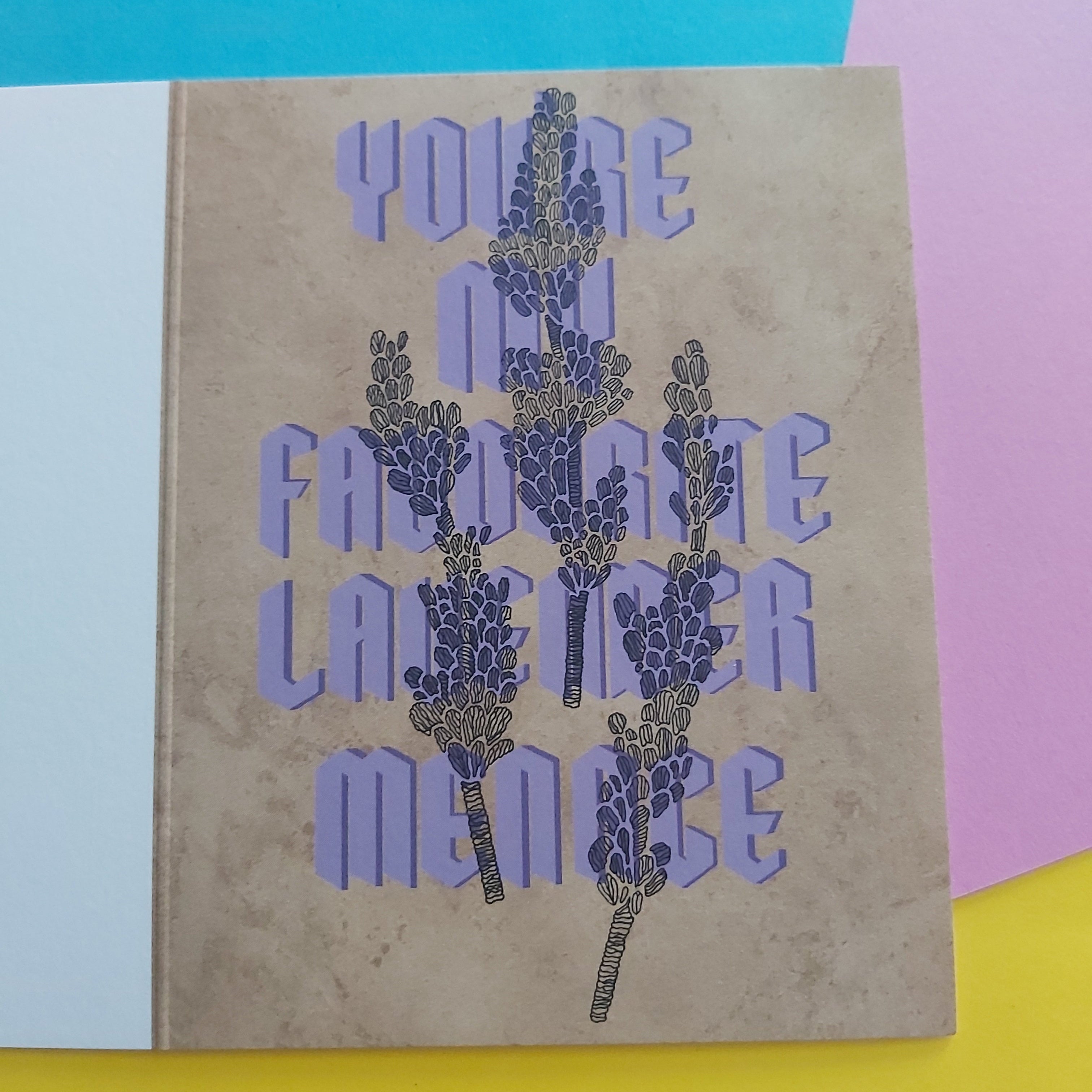 A greeting card with a textured beige background, featuring a purple and black lavender illustration and the text 'YOU'RE MY FAVORITE LAVENDER MENACE' on the front, displayed on a colourful background of yellow, pink, and blue paper.