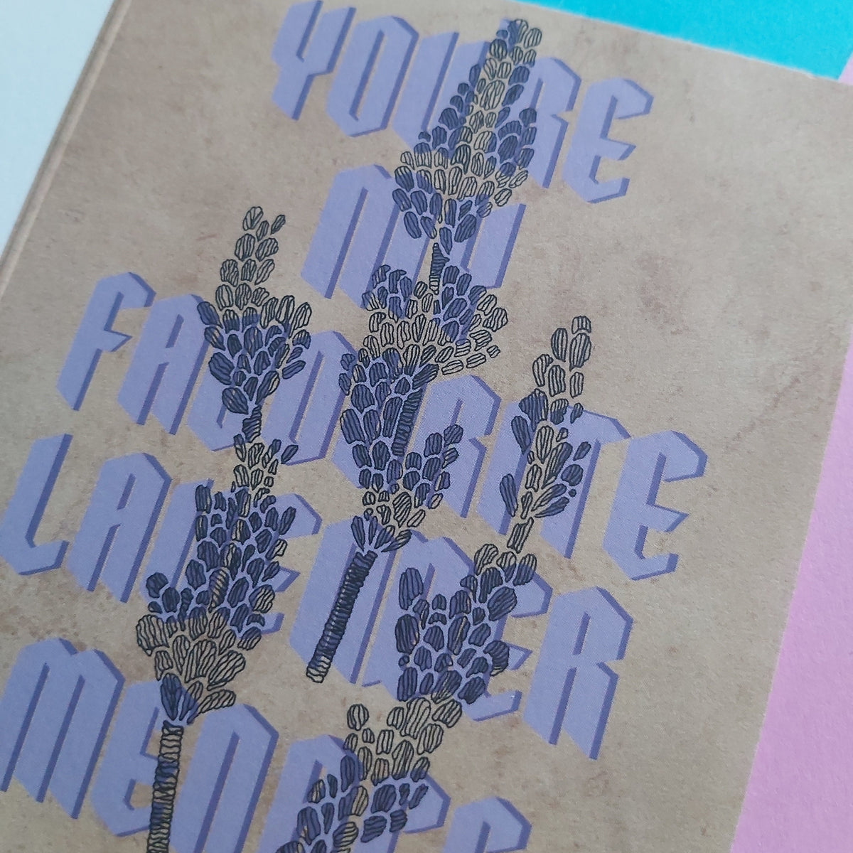 A close-up of a printed card page with the phrase 'YOU'RE MY FAVORITE LAVENDER MENACE' in large lavender-blue letters over a tan textured background, with illustrated lavender sprigs. A bright cyan corner and a pink corner are visible around the page.