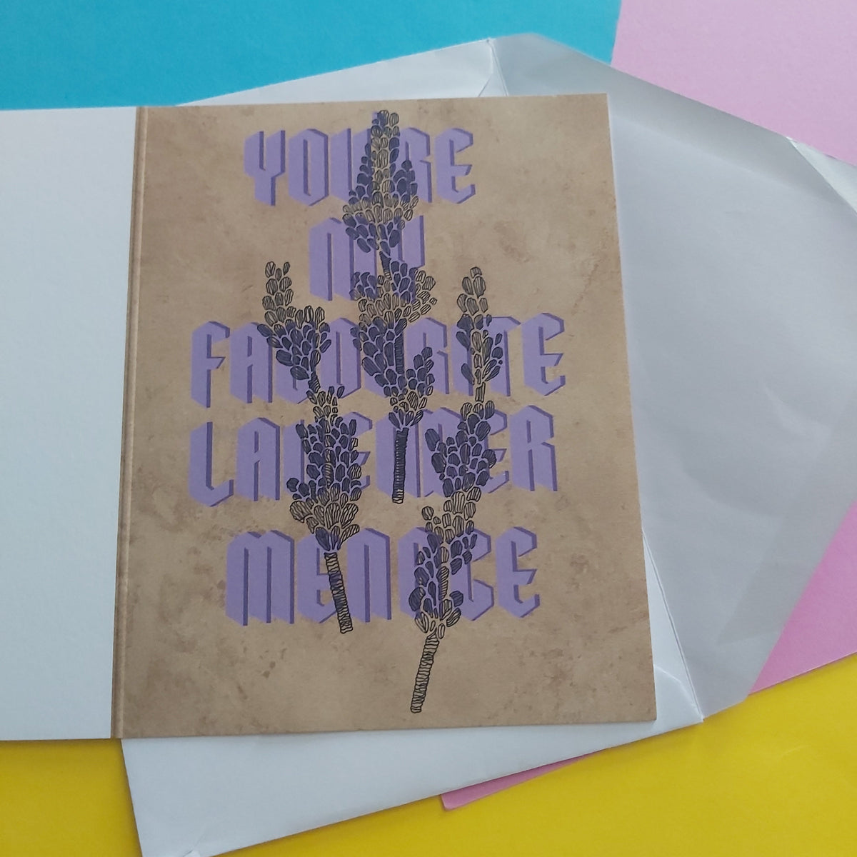 A greeting card with a textured tan background and purple text reading 'YOU'RE MY FAVORITE LAVENDER MENACE' overlaid with lavender illustrations, shown with a white envelope on a bright multicolour background.