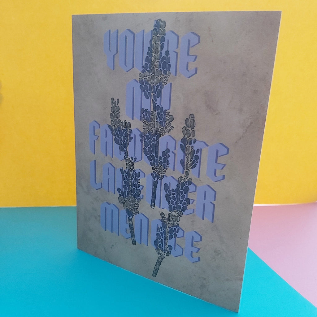 A greeting card with a textured beige background and a design featuring lavender flowers and the phrase 'YOU'RE MY FAVORITE LAVENDER MENACE' in purple letters, displayed on a colourful background of yellow, blue, and pink paper.