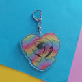 A heart-shaped acrylic keychain featuring a colorful rainbow gradient rose design with a silver lobster clasp, displayed on a bright blue background with colorful paper accents.