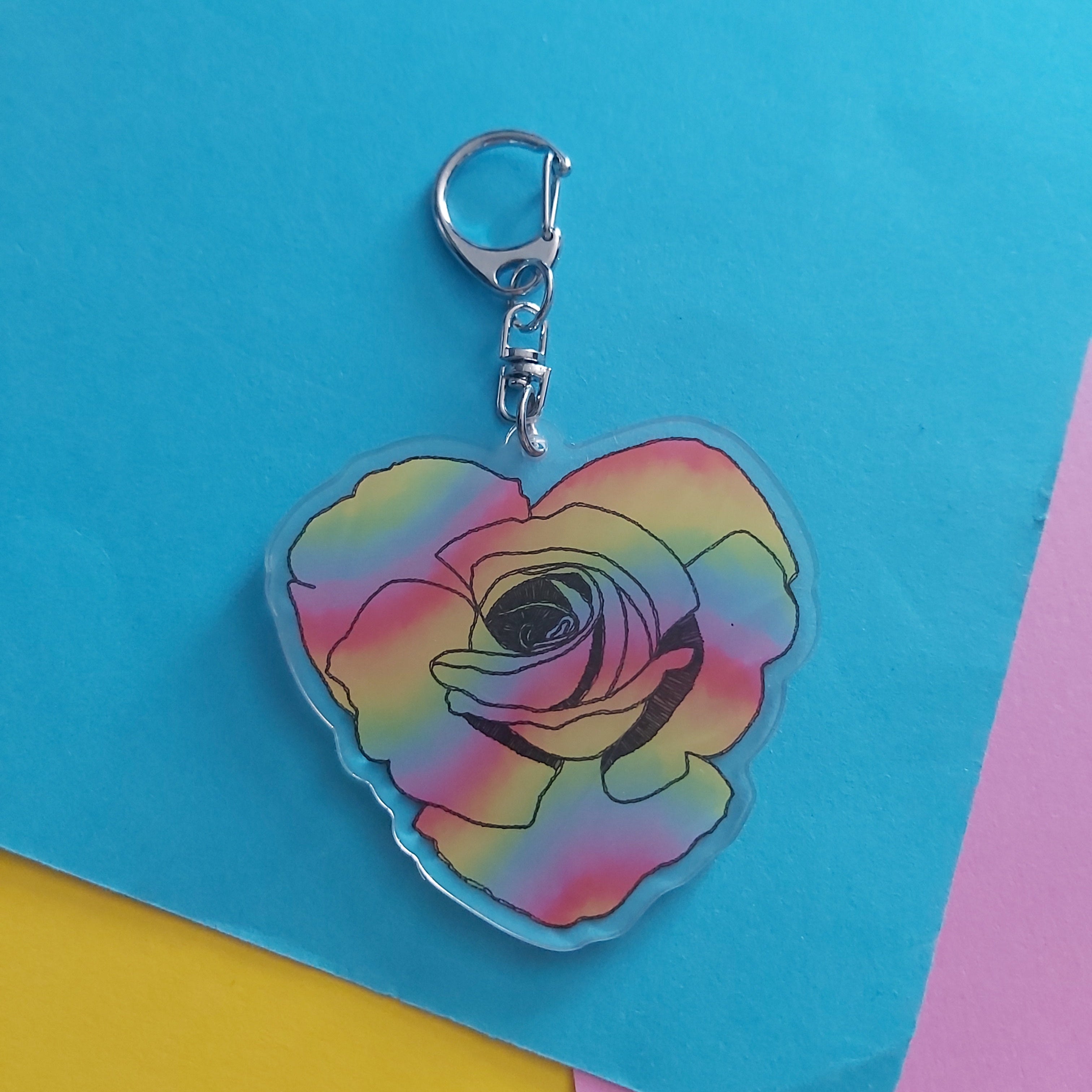 A heart-shaped acrylic keychain featuring a colorful rainbow gradient rose design with a silver lobster clasp, displayed on a bright blue background with colorful paper accents.