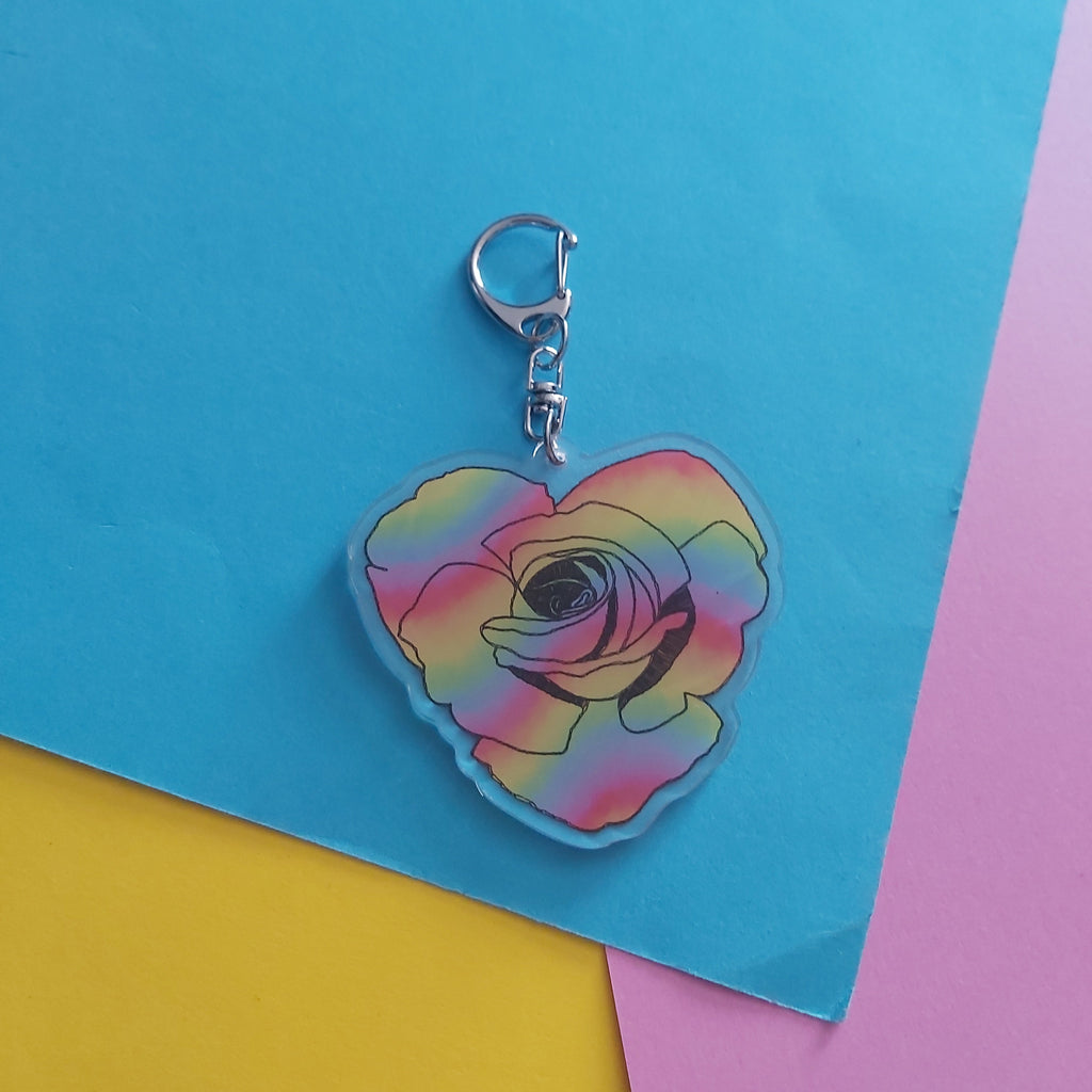 A heart-shaped acrylic keychain featuring a rose design with a rainbow gradient, complete with a silver metal clasp, set against a background of blue, yellow, and pink paper.