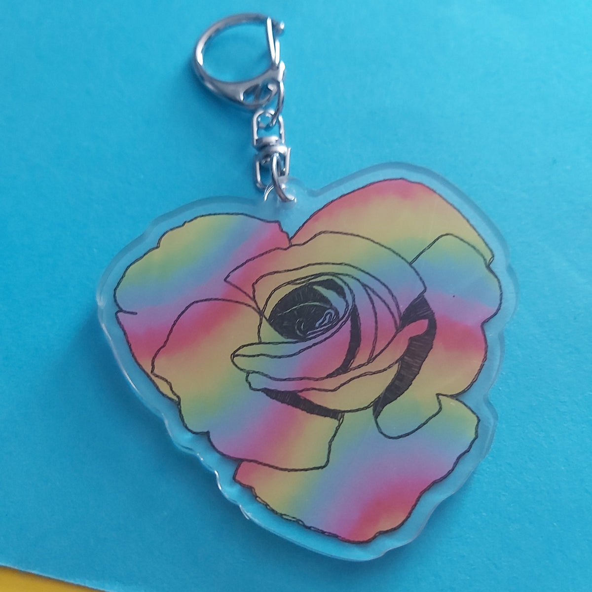 A heart-shaped acrylic keychain featuring a colorful rainbow gradient rose design with a silver lobster clasp, displayed on a bright blue background with yellow and pink paper corners visible.
