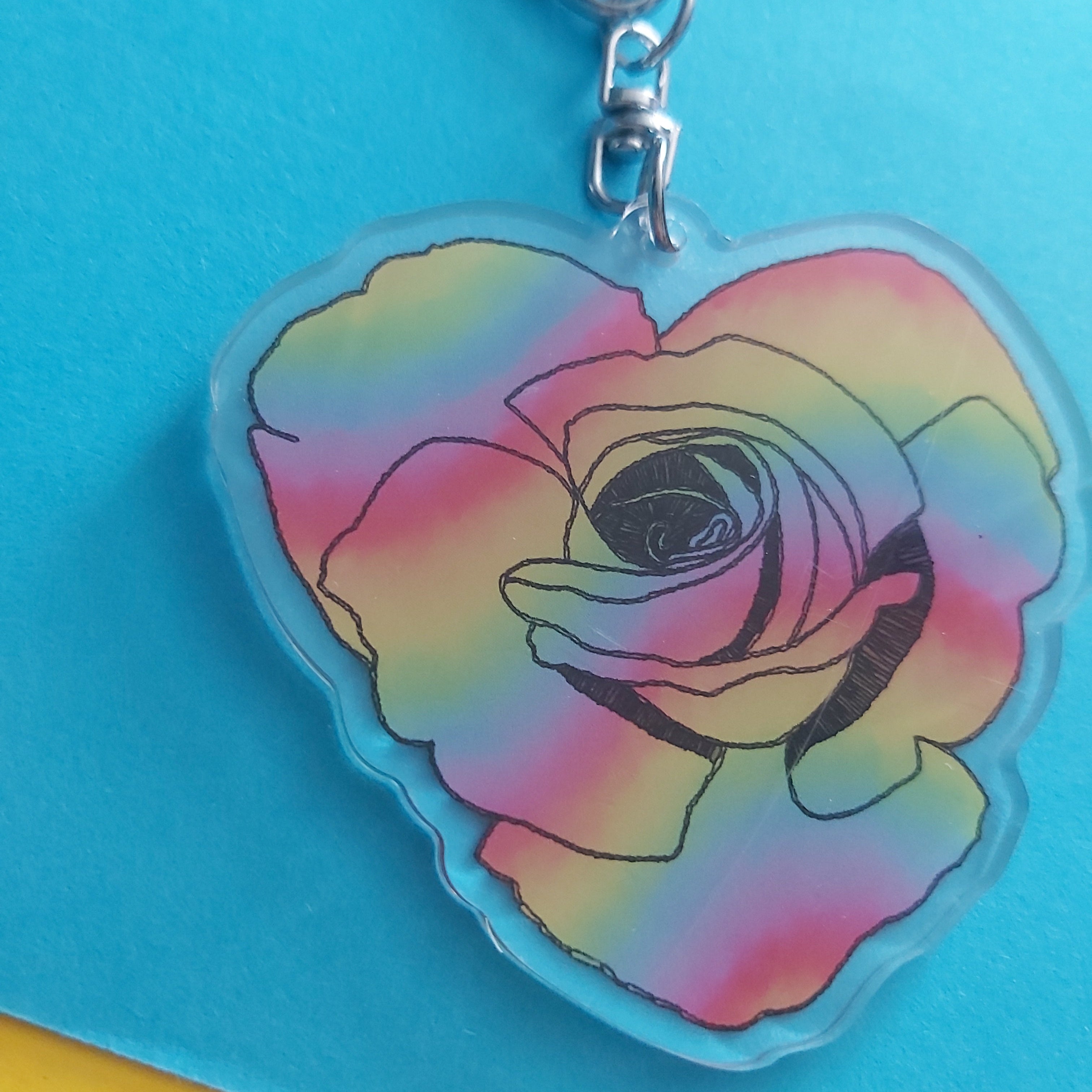 A heart-shaped acrylic keychain featuring a rose design with a rainbow gradient of pink, yellow, green, and blue, attached to a silver metal clasp, set against a bright blue background.