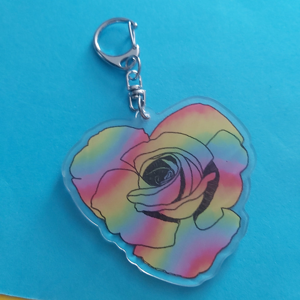 A heart-shaped acrylic keychain featuring a rose design with a rainbow gradient of pink, yellow, green, and blue, attached to a silver lobster clasp, set against a bright blue background.