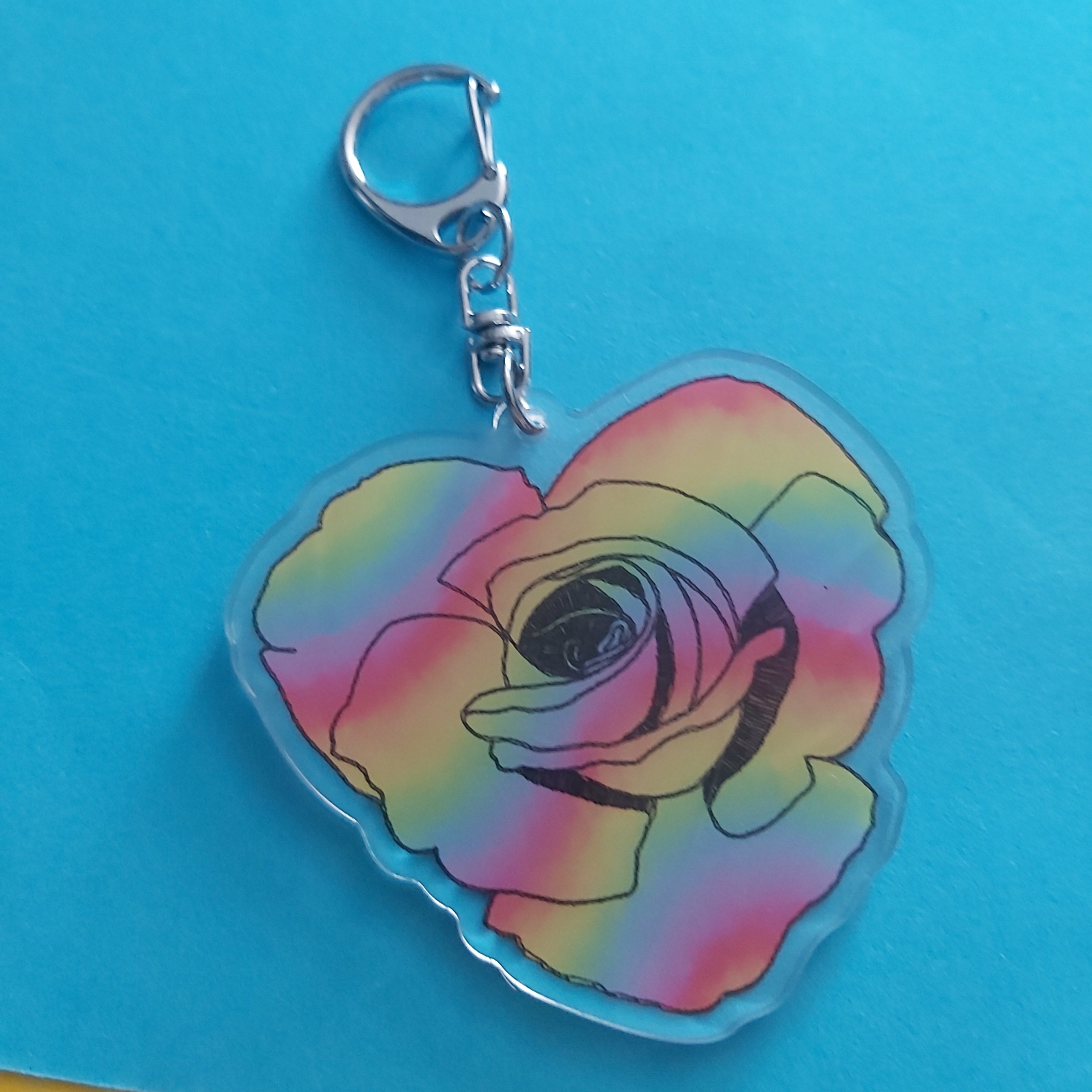 A heart-shaped acrylic keychain featuring a rose design with a rainbow gradient of pink, yellow, green, and blue, attached to a silver lobster clasp, set against a bright blue background.