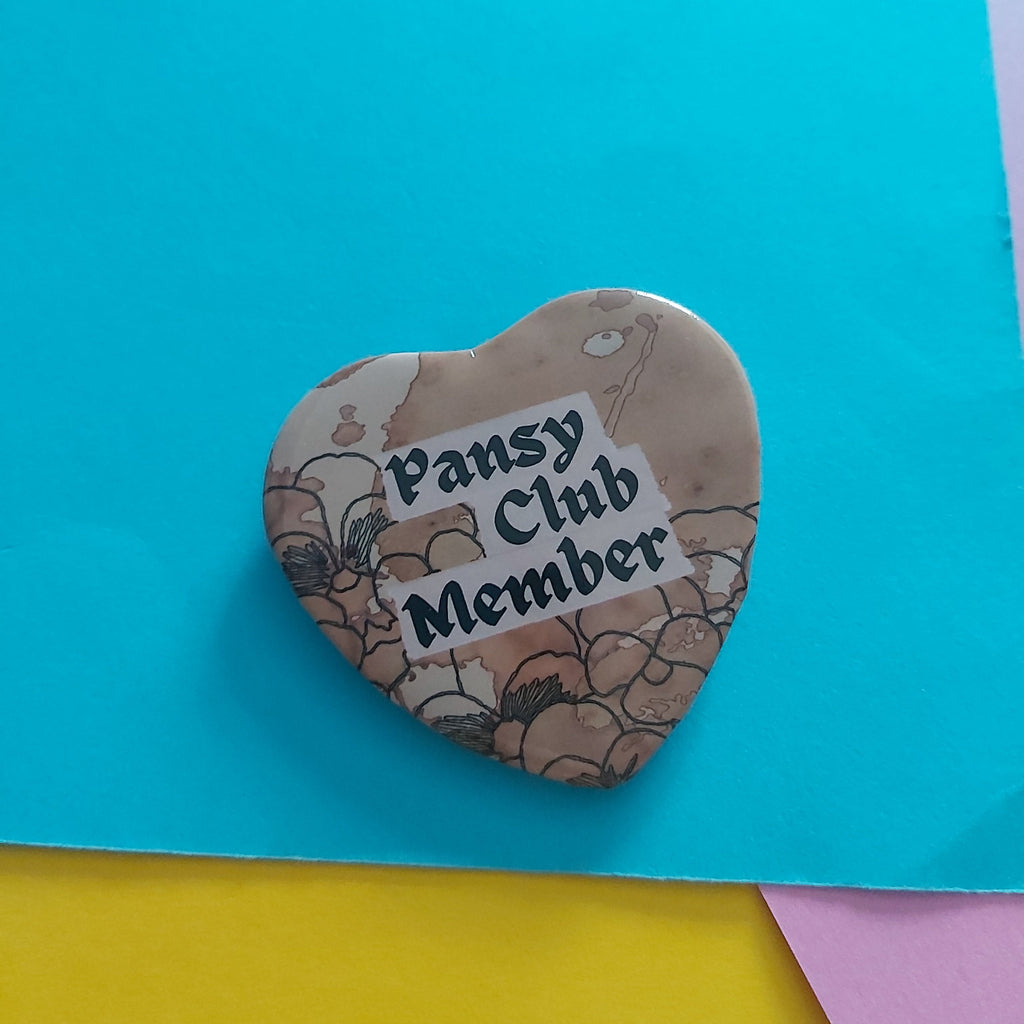A heart-shaped button badge with a floral pattern and the text 'Pansy Club Member' in black lettering on a white label, placed on a bright blue background with yellow and pink paper corners visible.
