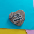 A heart-shaped button badge with a floral pattern and the text 'Pansy Club Member' in black lettering on a white label, placed on a bright blue background with yellow and pink paper corners visible.