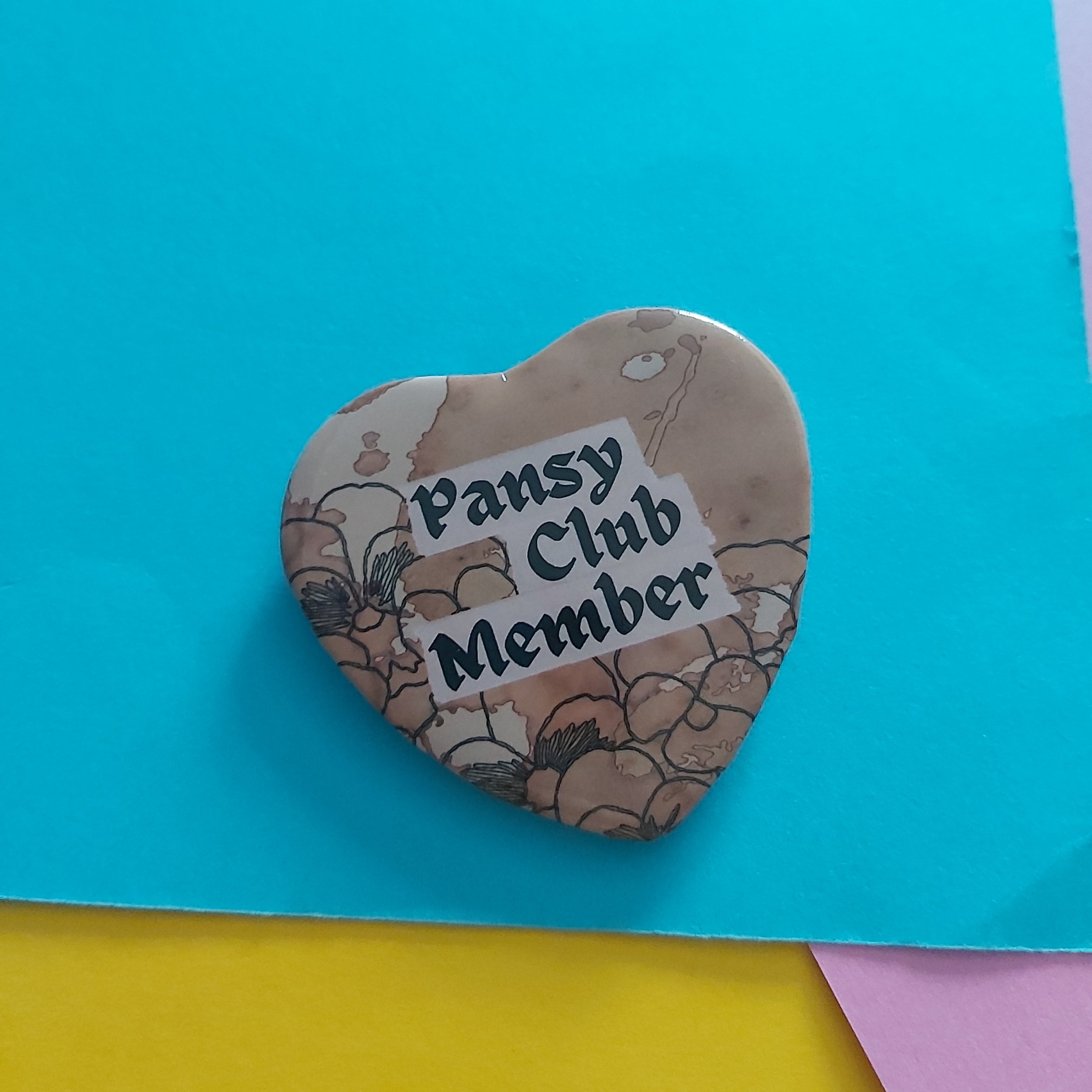 A heart-shaped button badge with a floral pattern and the text 'Pansy Club Member' in black lettering on a white label, placed on a bright blue background with yellow and pink paper corners visible.