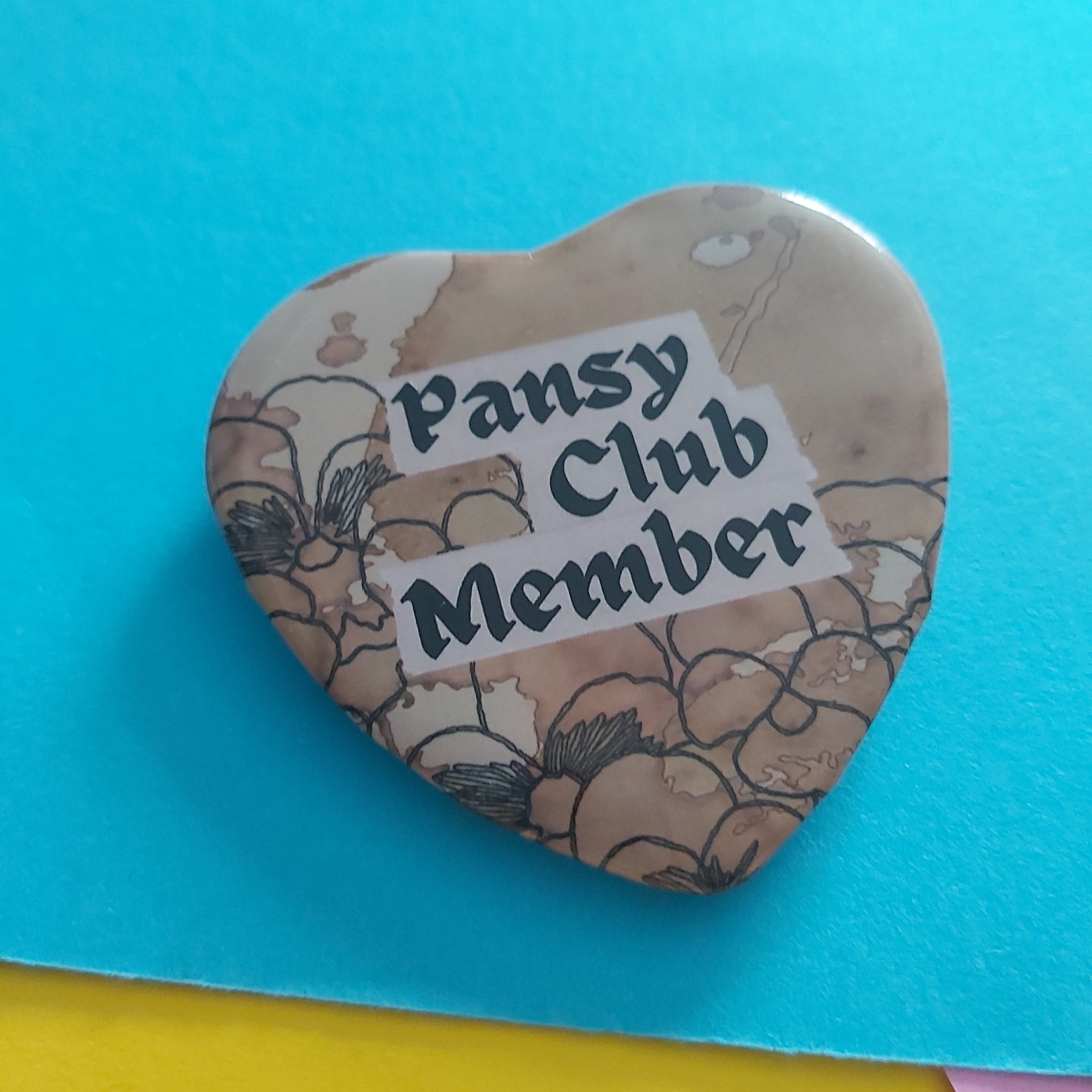 A heart-shaped pin badge with a vintage floral pattern in beige and brown, featuring a white banner that reads 'Pansy Club Member' in black text, set against a bright blue background with yellow and pink paper accents.