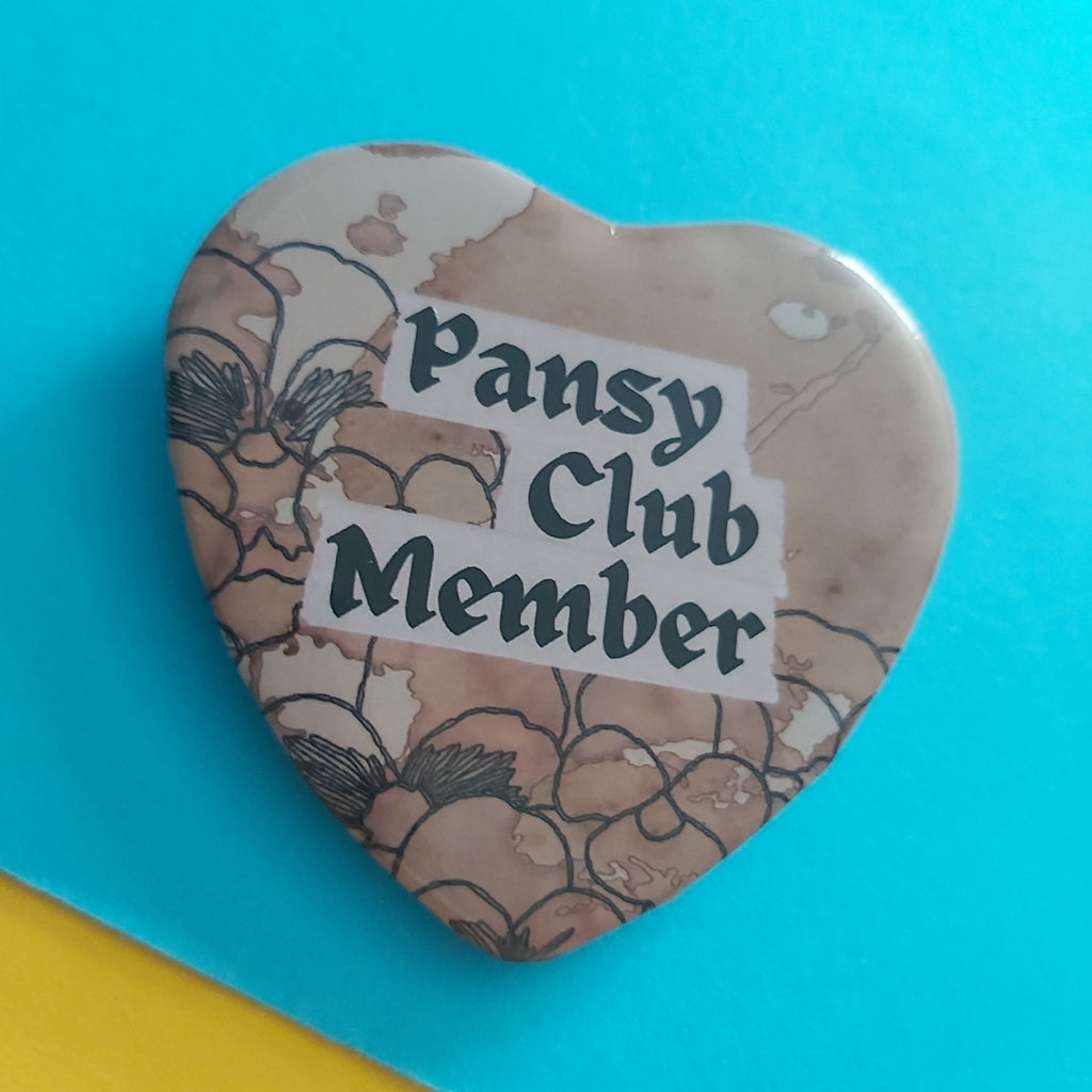 A heart-shaped pin badge with a beige and brown floral pattern, featuring a white banner that reads 'Pansy Club Member' in black text, set against a bright blue background with a yellow corner visible.