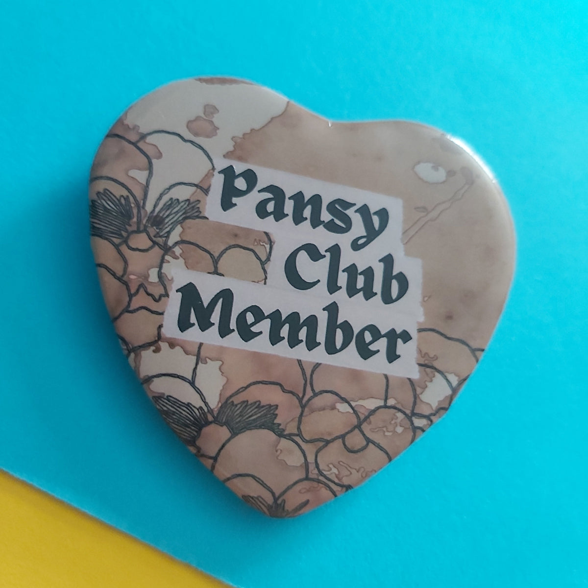 A heart-shaped pin badge with a beige and brown floral pattern, featuring a white banner that reads 'Pansy Club Member' in black text, set against a bright blue background with a yellow corner visible.