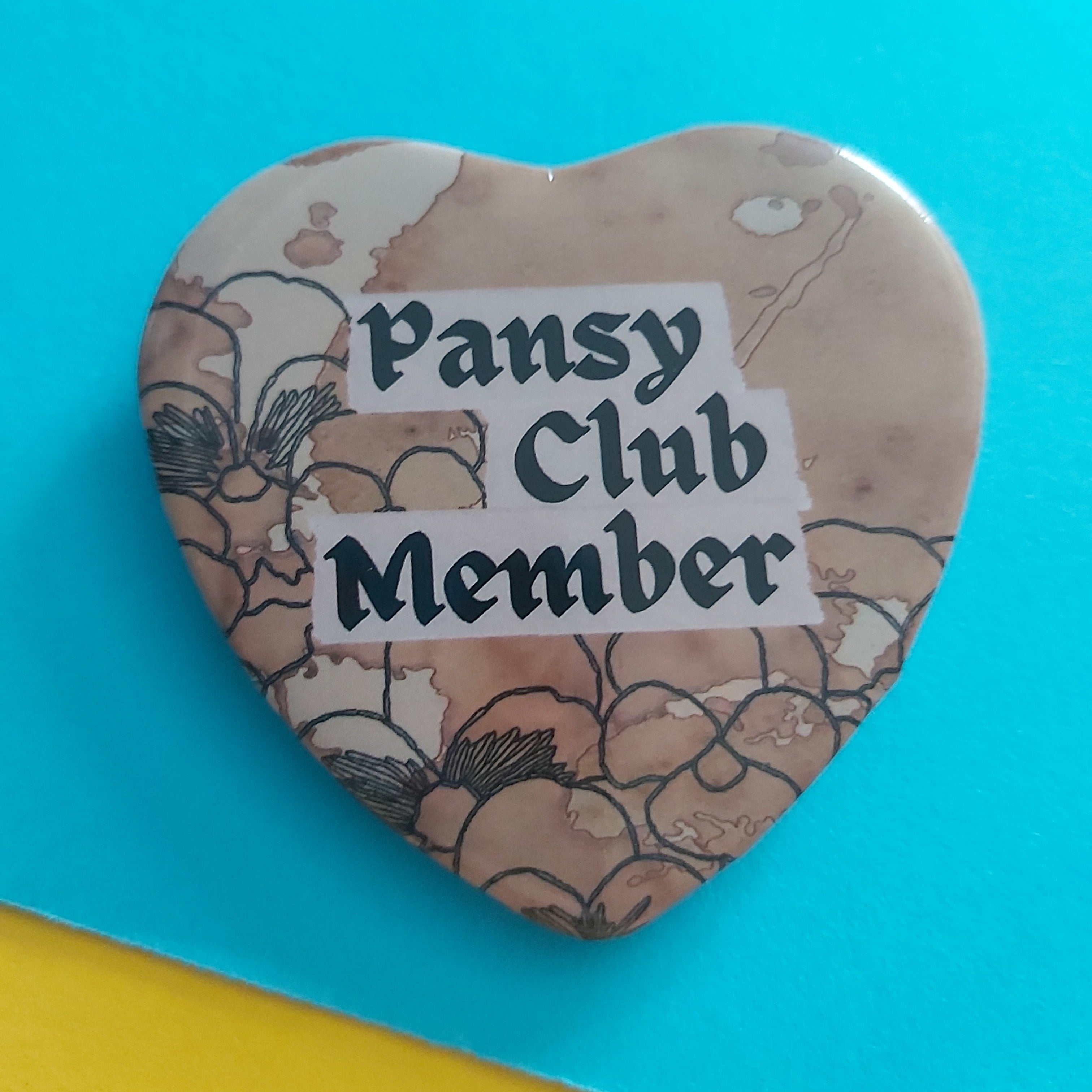 A heart-shaped button badge featuring a vintage-style floral pattern in beige and brown tones, with a white label reading 'Pansy Club Member' in black gothic lettering, set against a bright turquoise background.