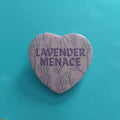 A heart-shaped pinback button badge featuring the words 'LAVENDER MENACE' in purple text over a lavender-purple background with line-art illustrations of lavender plants. The badge is placed on a bright turquoise paper background.