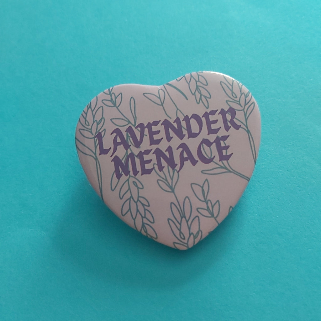 A heart-shaped pin badge featuring the words 'LAVENDER MENACE' in purple text surrounded by lavender illustrations, set against a bright turquoise background.