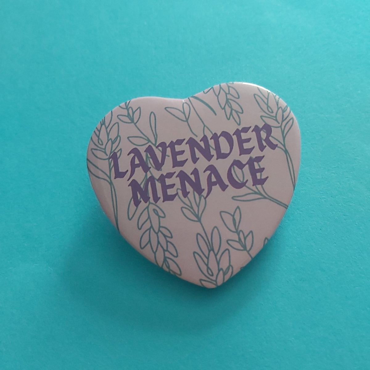 A heart-shaped pin badge featuring the words 'LAVENDER MENACE' in purple text surrounded by lavender illustrations, set against a bright turquoise background.