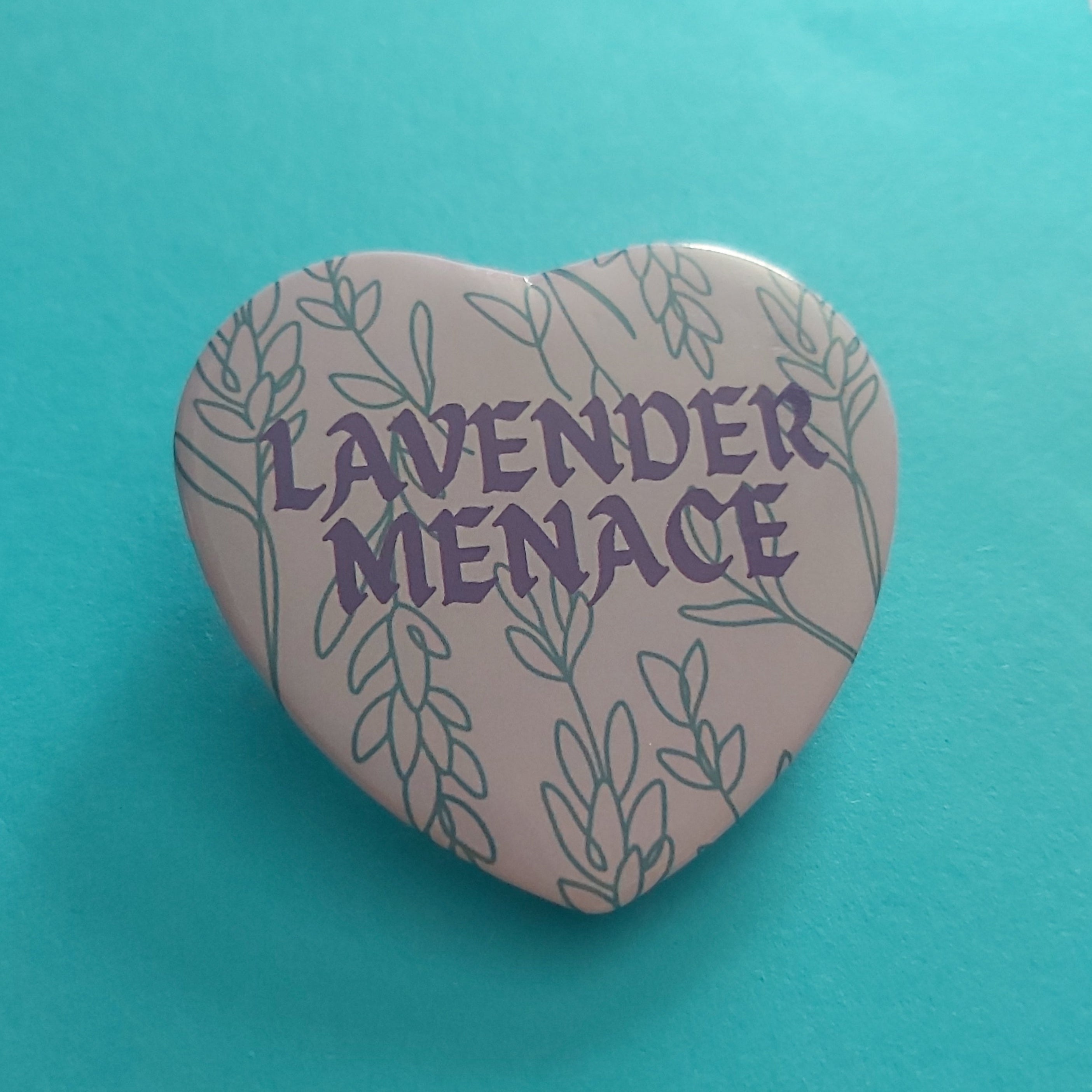 A heart-shaped pin badge featuring the text 'LAVENDER MENACE' in purple, surrounded by a lavender leaf pattern on a light purple background, set against a bright turquoise paper backdrop.