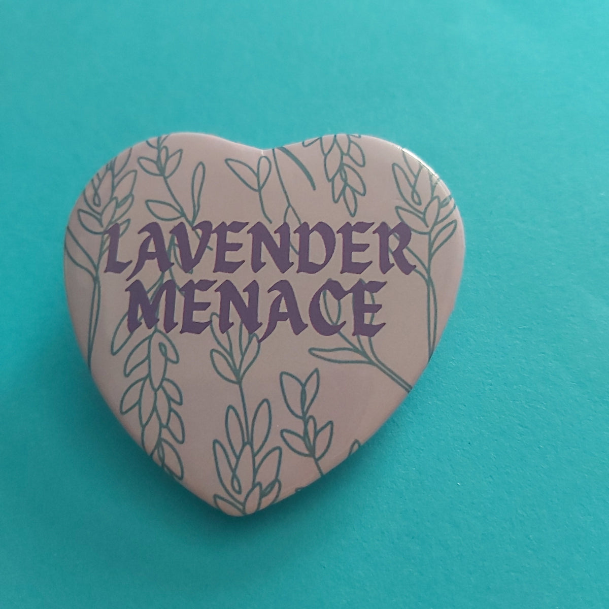 A heart-shaped pin badge featuring the text 'LAVENDER MENACE' in purple lettering, surrounded by a lavender botanical line pattern on a pale purple background, set against a solid teal background.