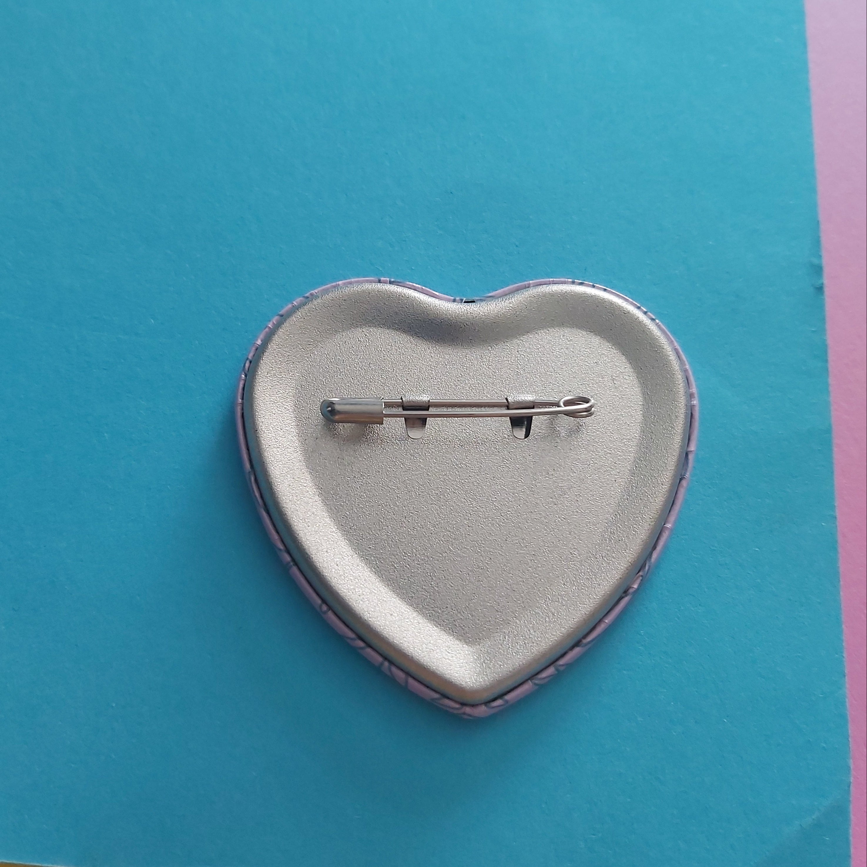 A heart-shaped pinback button showing its silver metal back with a safety pin clasp, resting on a bright blue sheet of paper with pink paper edges visible.
