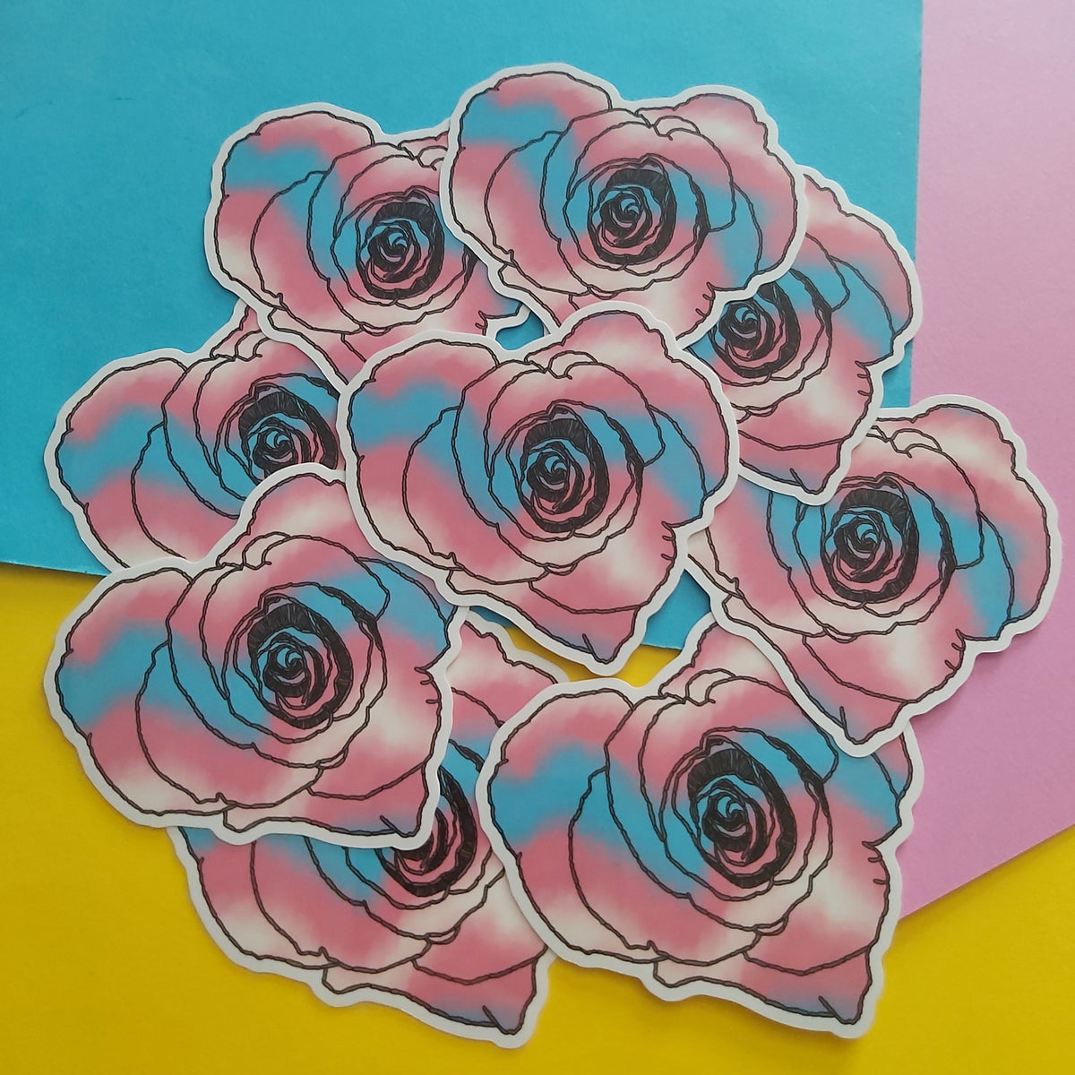 A cluster of die-cut stickers featuring stylised pink and blue roses with black outlines, arranged on a background of bright yellow, pink, and blue paper.