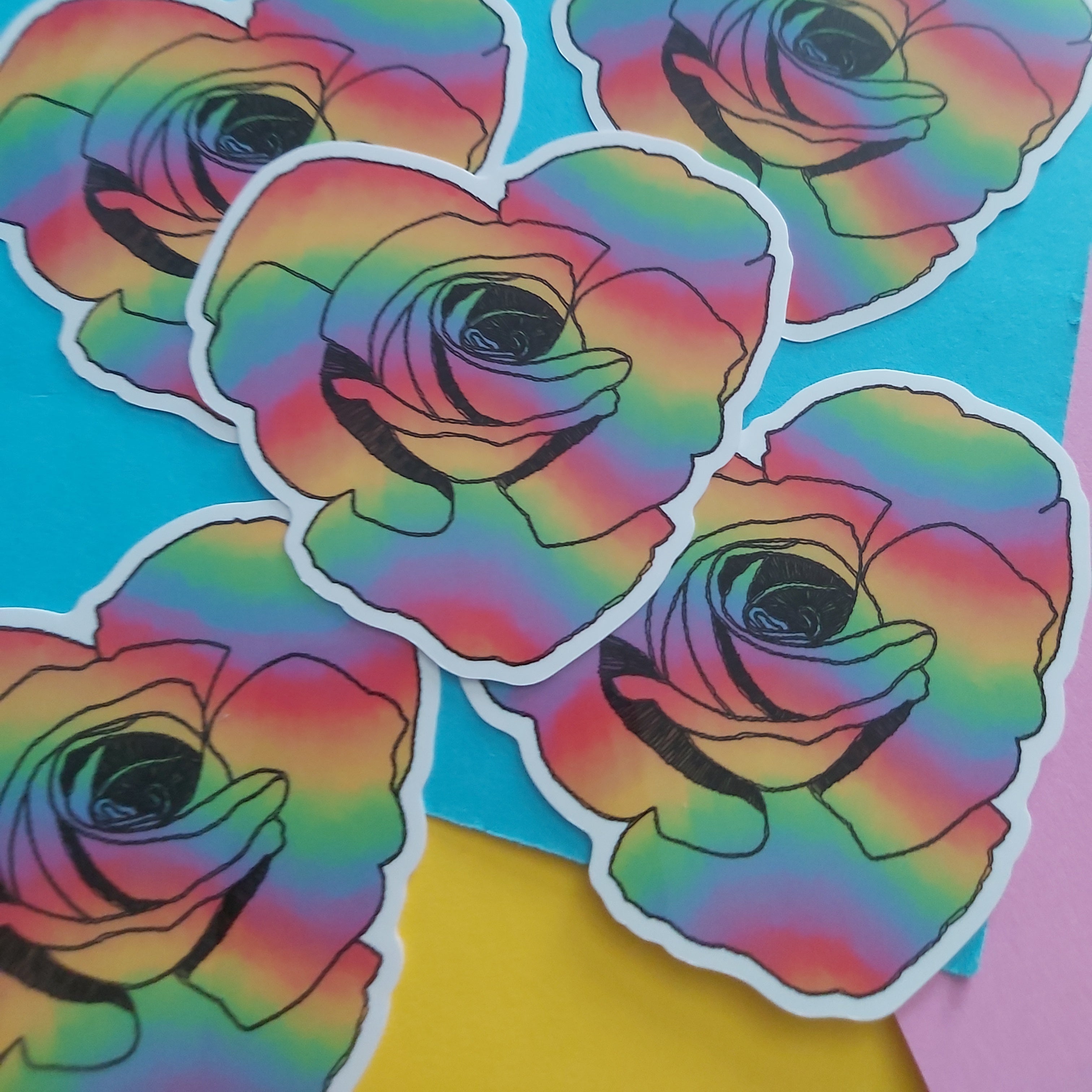 Five die-cut stickers featuring a rainbow-striped rose design with black outlines and white borders, arranged on a bright blue background with yellow and pink paper accents.
