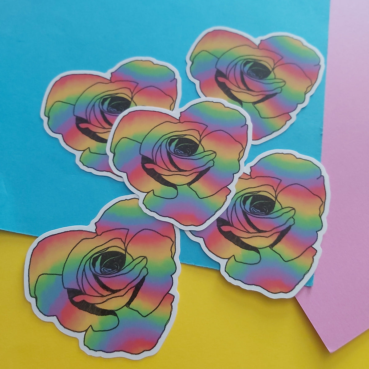 Five die-cut stickers featuring a black line art rose filled with a rainbow gradient, arranged on a split yellow and blue background with a pink paper corner.