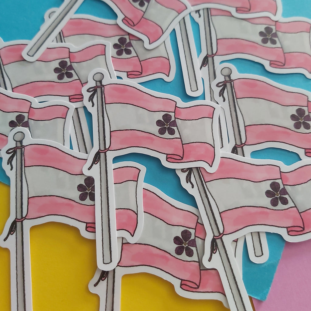 A pile of die-cut stickers depicting the sapphic flag, featuring pink and white horizontal stripes with a floral symbol in the centre, mounted on silver flagpoles, set against a colourful background of blue, yellow, and pink paper.