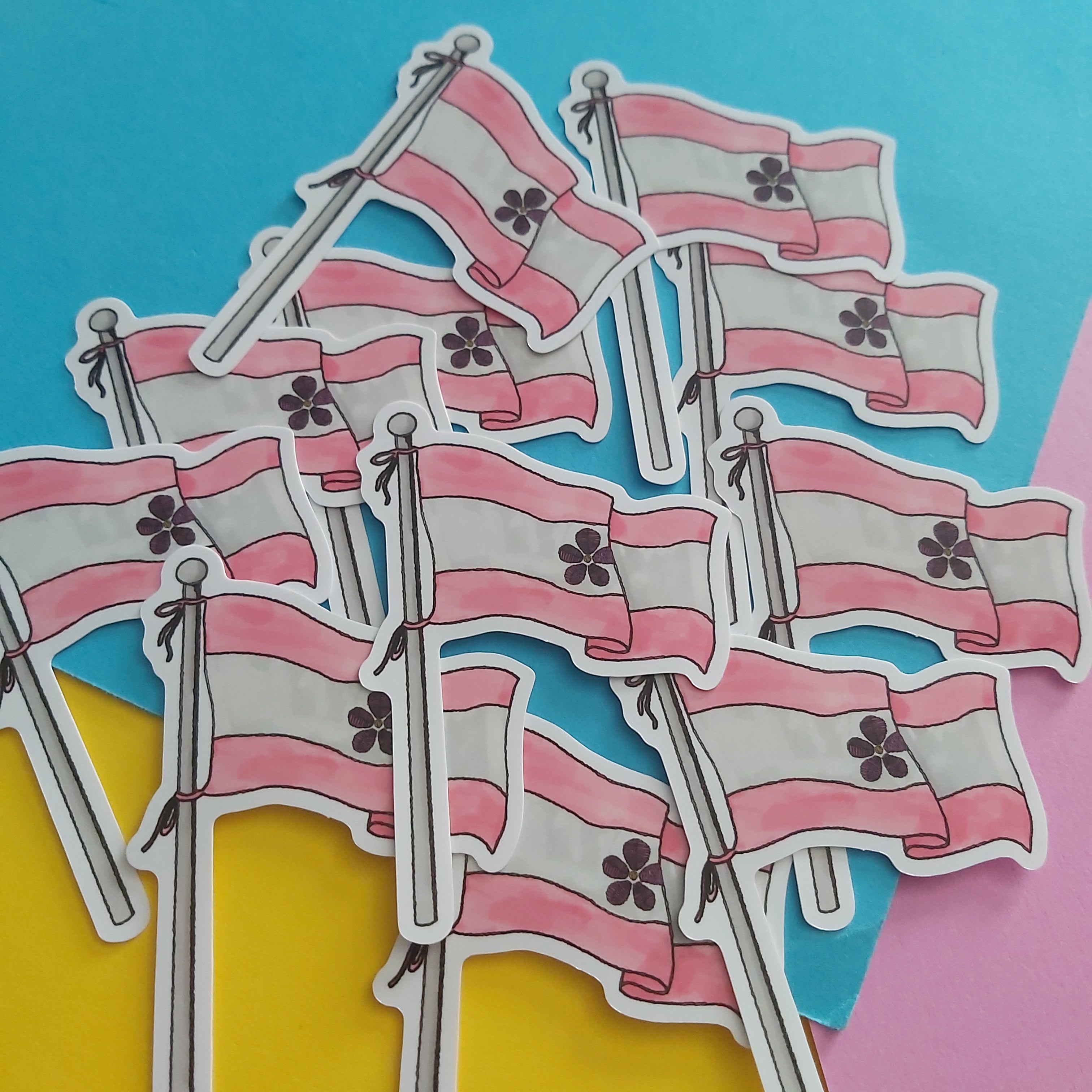 A pile of small flag stickers with pink and white horizontal stripes and a dark flower emblem in the center, arranged on a bright blue, yellow, and pink background.