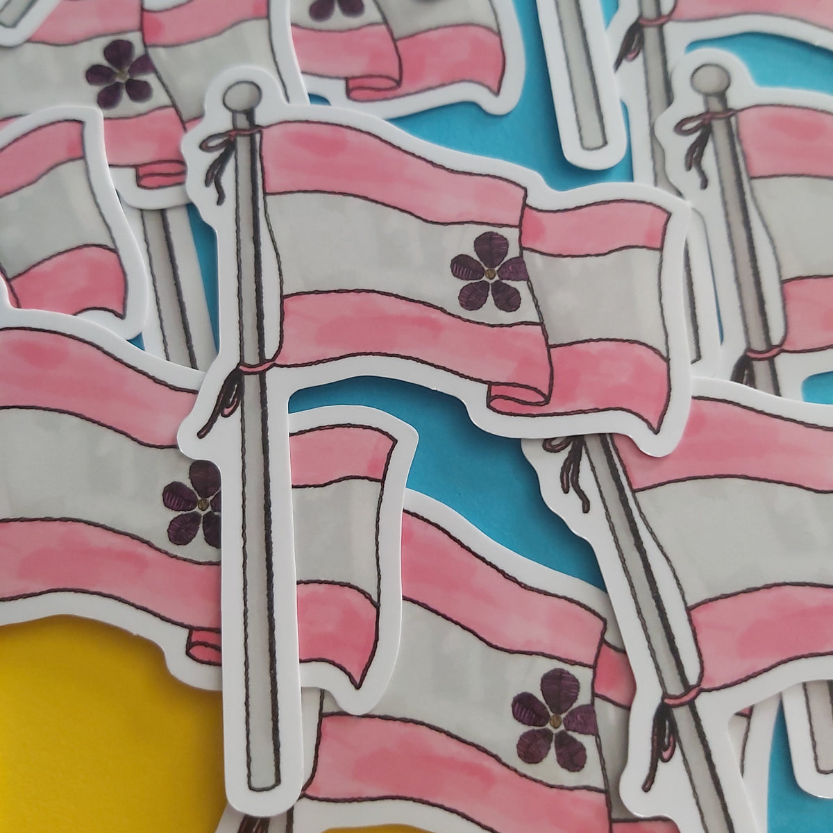 A close-up of multiple die-cut stickers depicting the sapphic pride flag on poles, arranged overlapping on a bright blue and yellow background.