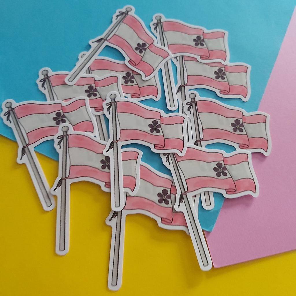 A pile of small die-cut stickers depicting the sapphic pride flag with pink and white horizontal stripes and a black-outlined floral emblem, arranged on a colourful background of yellow, blue, and pink paper.