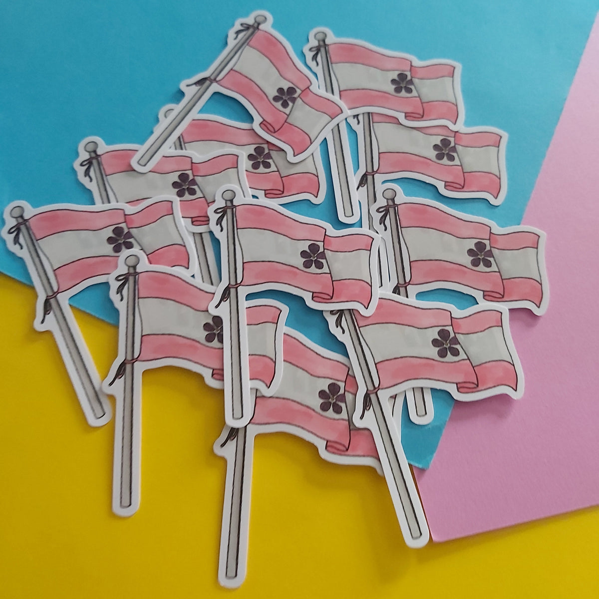 A pile of small die-cut stickers depicting the sapphic pride flag with pink and white horizontal stripes and a black-outlined floral emblem, arranged on a colourful background of yellow, blue, and pink paper.