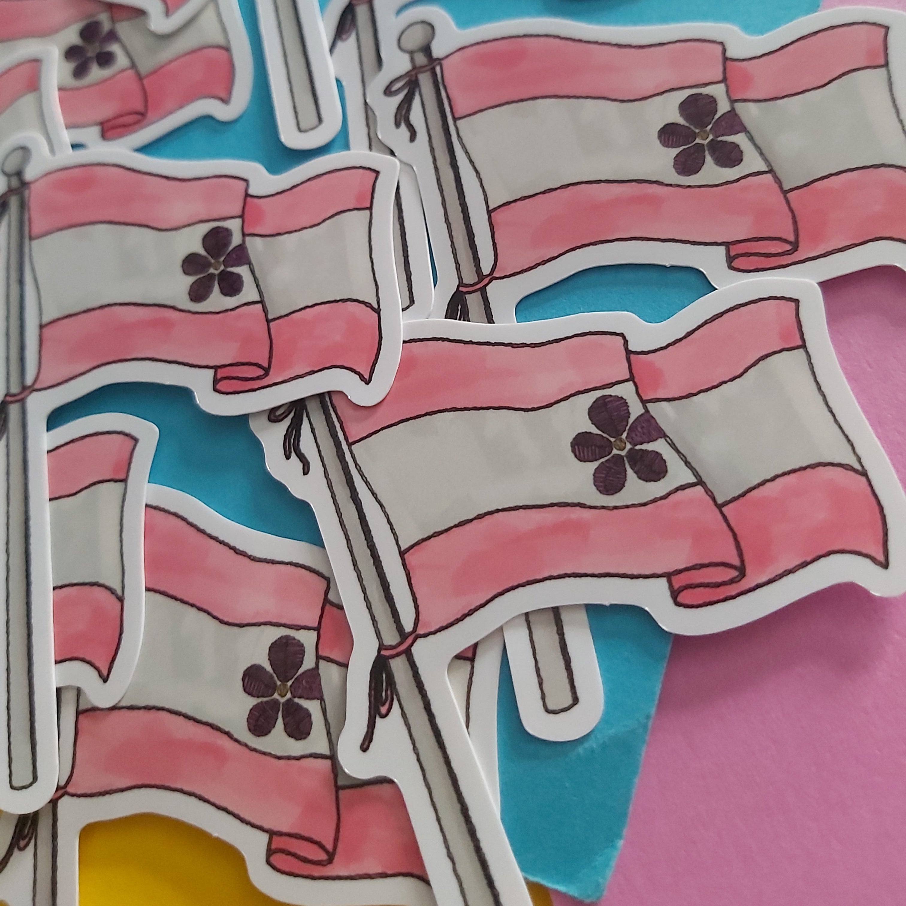 A pile of die-cut stickers depicting the sapphic flag, featuring pink and white horizontal stripes with a flower emblem and a grey flagpole, set against a colourful background of blue, pink, and yellow paper.