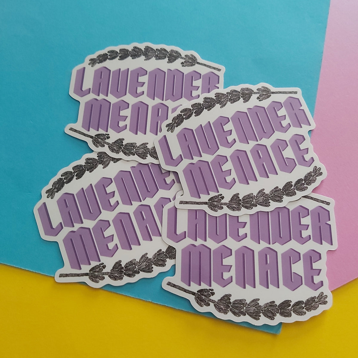 Four die-cut stickers featuring the phrase 'LAVENDER MENACE' in purple retro-style lettering with a black floral border, arranged on a colourful background of blue, pink, and yellow paper.