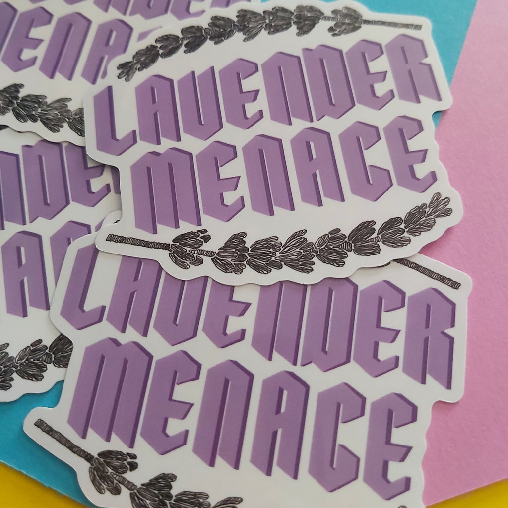 A stack of die-cut stickers featuring the words 'LAVENDER MENACE' in purple block letters with a dark lavender branch illustration, set against a background of blue, pink, and yellow paper.