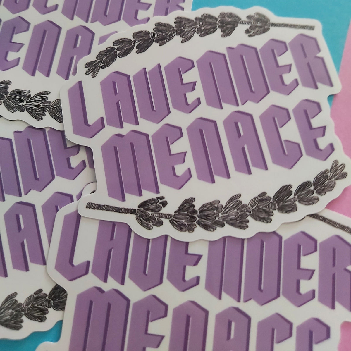 A close-up of several die-cut stickers featuring the words 'LAVENDER MENACE' in purple block letters with a black lavender sprig graphic, arranged on a bright blue background.
