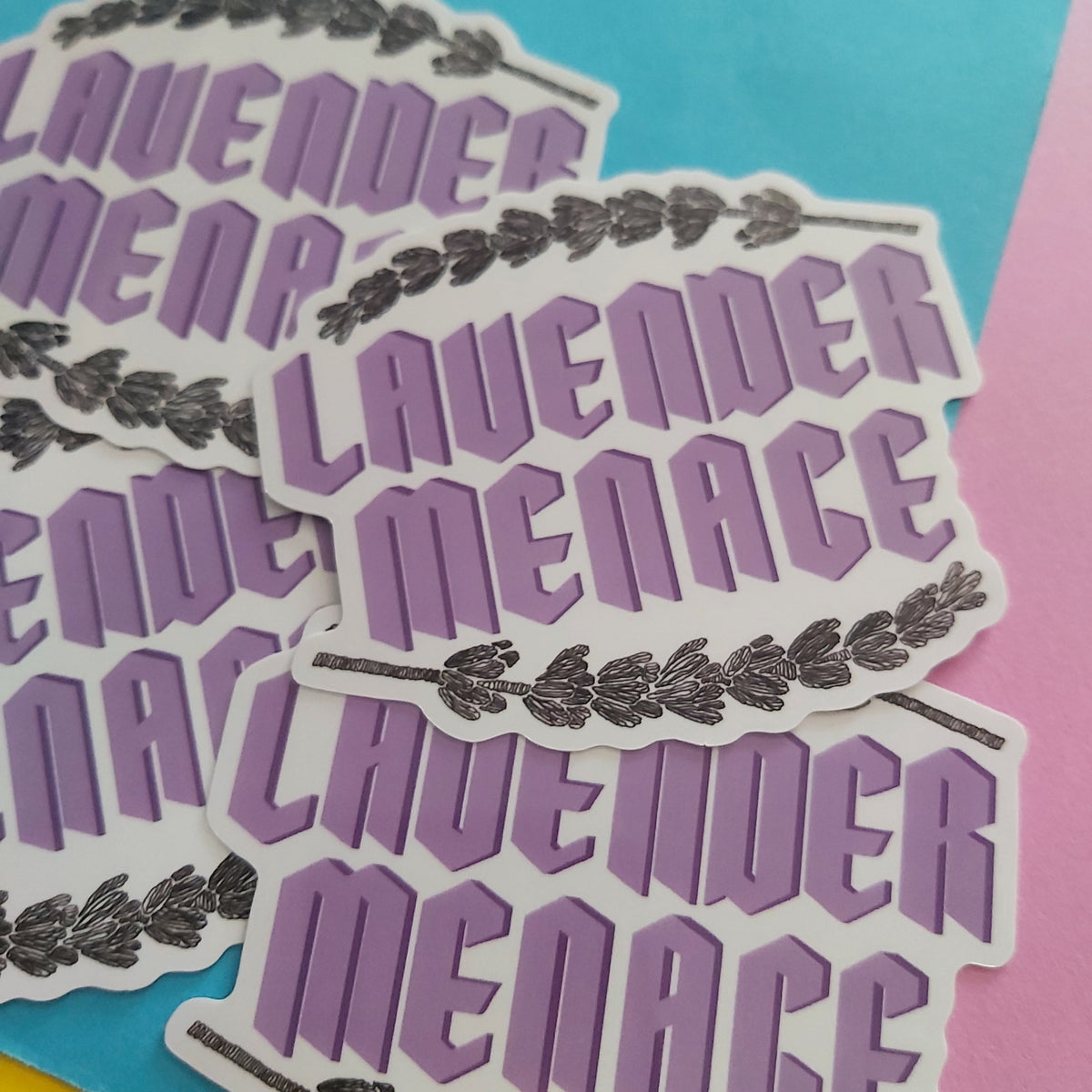 Several die-cut stickers reading “LAVENDER MENACE” in lavender-purple lettering with a leafy border, arranged on a bright blue background with pink and yellow sections visible.