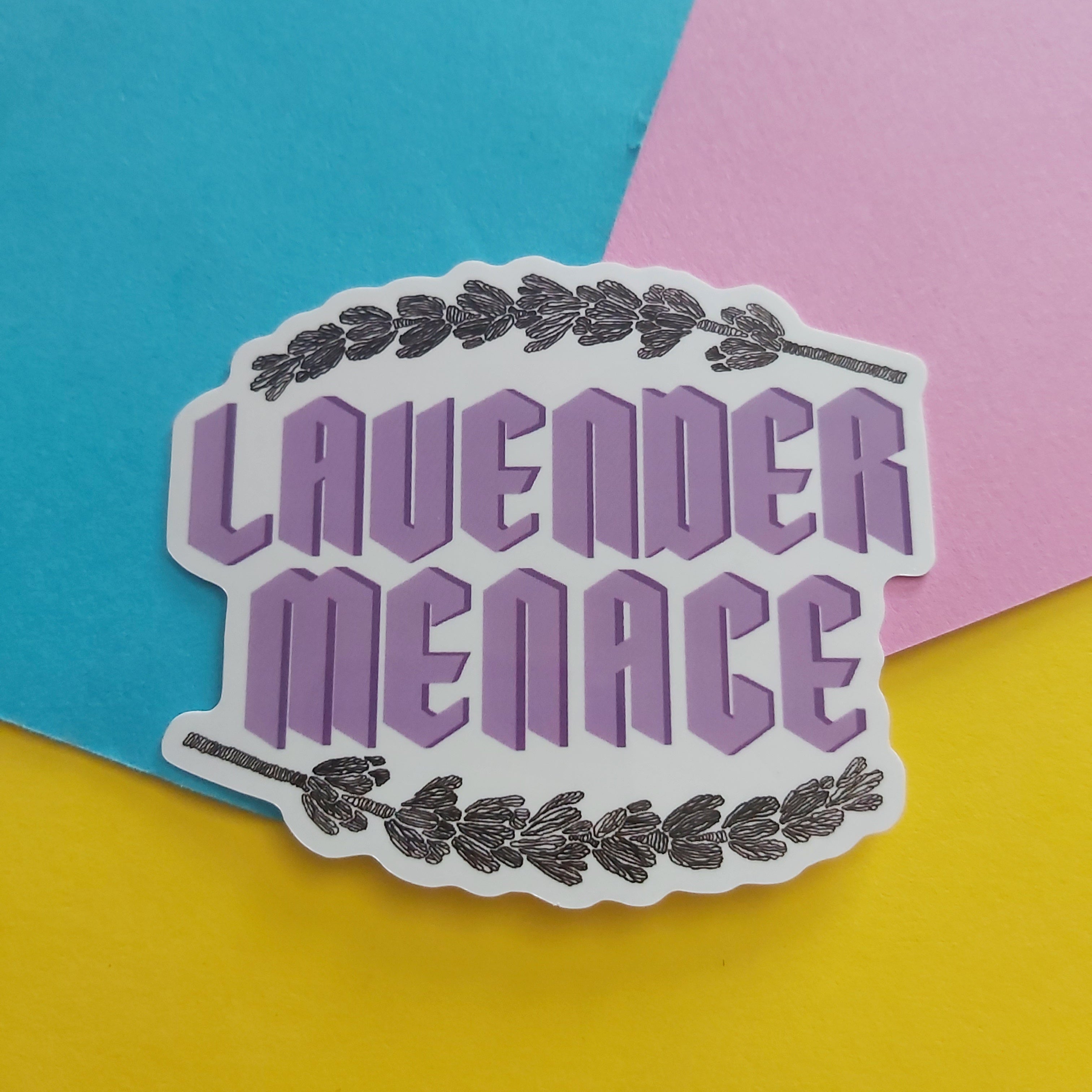A die-cut sticker with the words 'LAVENDER MENACE' in lavender-purple block letters, framed by dark lavender sprigs at the top and bottom, shown on a colourful background of blue, pink, and yellow paper.