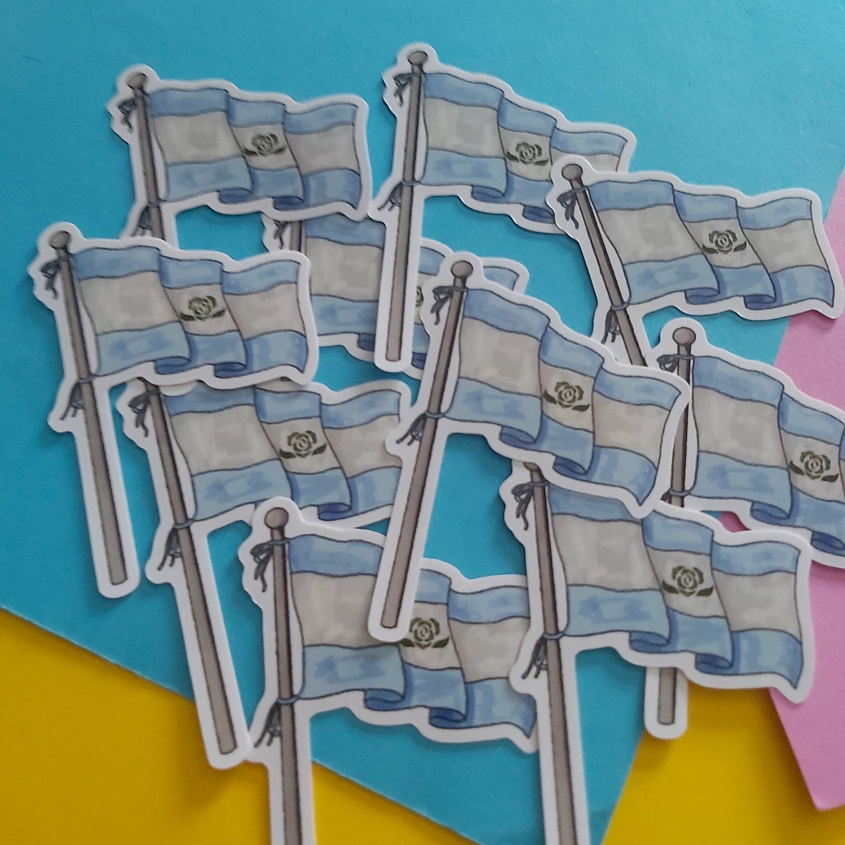 A pile of die-cut stickers depicting the Achillean pride flag on a colourful background of blue, yellow, and pink paper