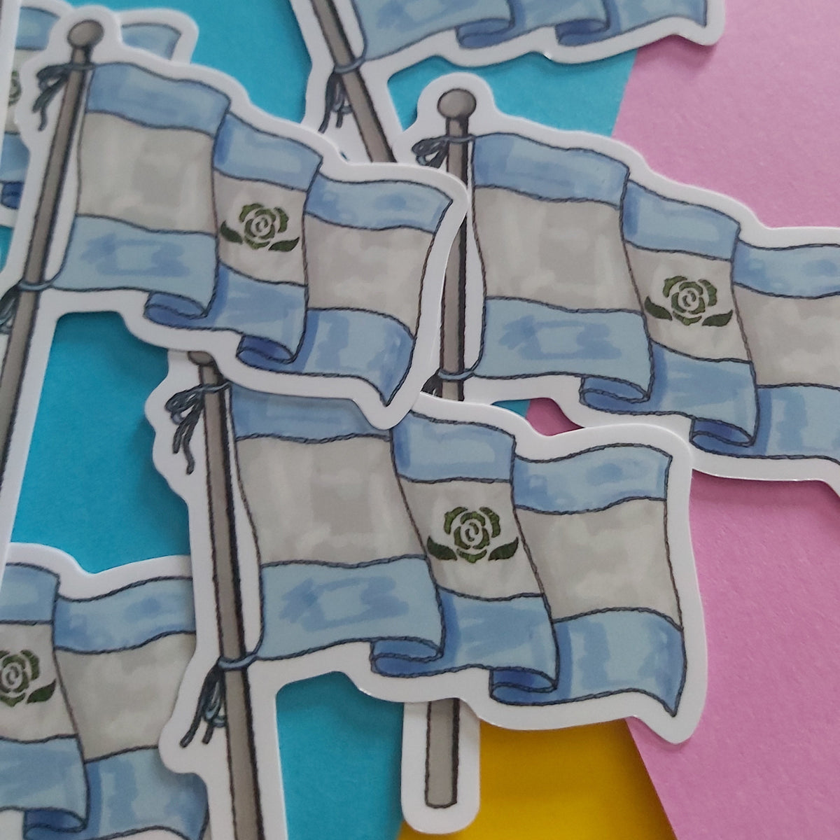 A close-up of several die-cut stickers depicting the Achillean flag on poles, arranged on a colorful background with cyan, pink, and yellow paper.