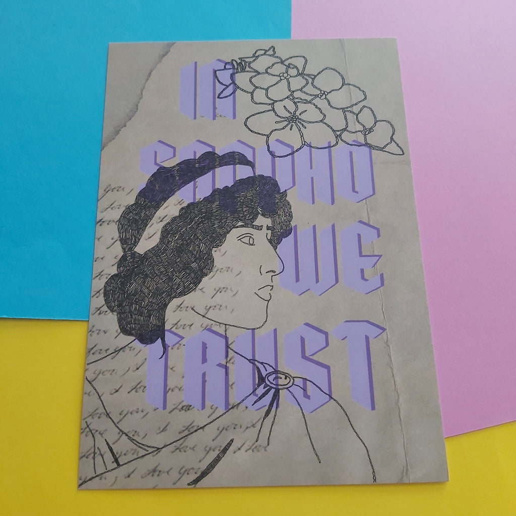An art print featuring a vintage-style illustration of a woman in profile with flowers above her head, overlaid with large lavender text reading 'IN SAPHO WE TRUST' on a beige background. The print is photographed on a bright yellow surface with blue and pink paper shapes behind it.