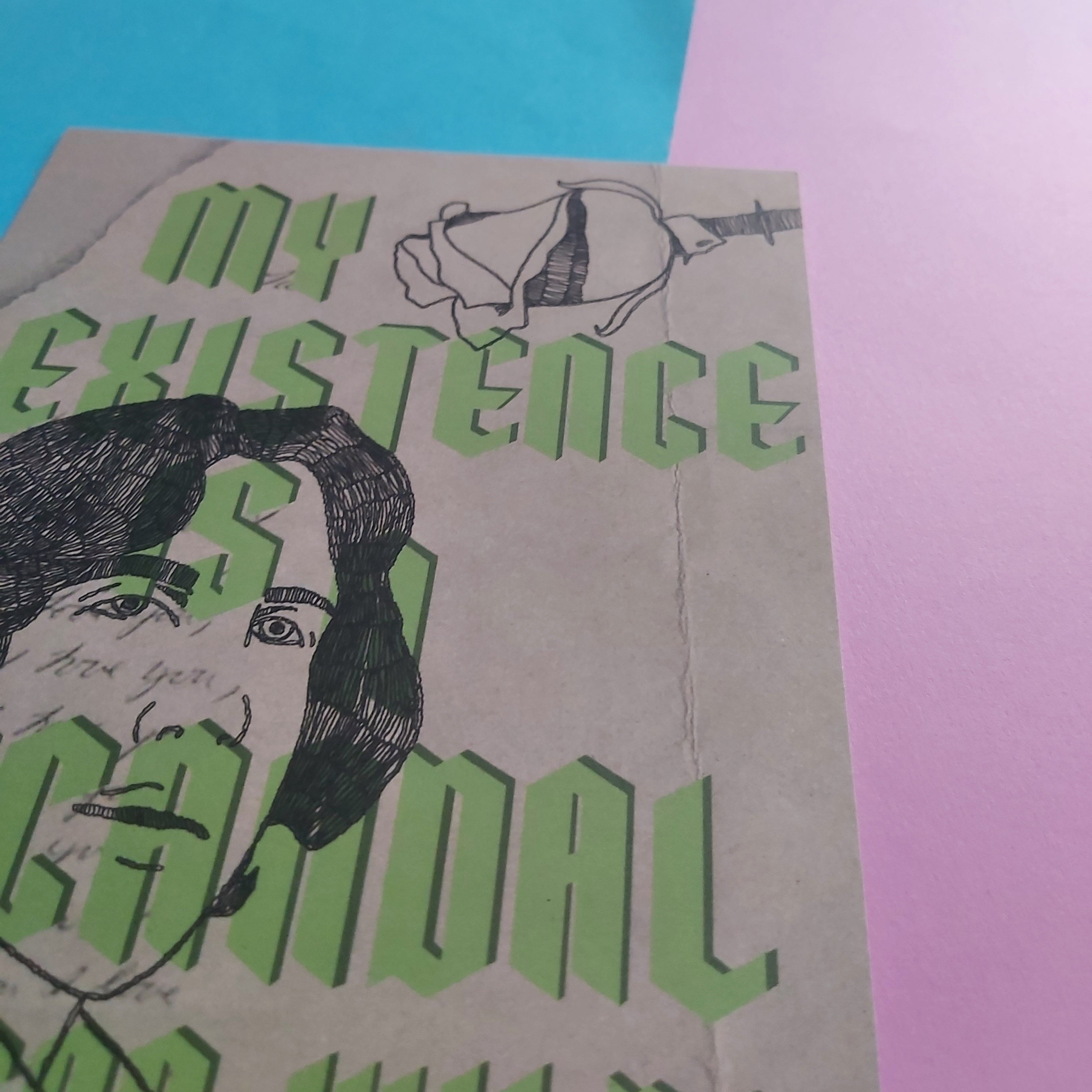 A close-up of a print with the phrase 'MY EXISTENCE IS A SCANDAL' in large green gothic-style letters over a black line drawing of a face and a flower, placed on a surface with blue and pink paper behind it.