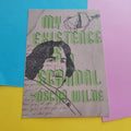A print featuring a stylised portrait of Oscar Wilde with the text 'MY EXISTENCE IS A SCANDAL -OSCAR WILDE' in green, layered over sketchy black line art and cursive script on a beige background. The card is photographed on a bright yellow surface with blue and pink paper sheets partially visible behind it.