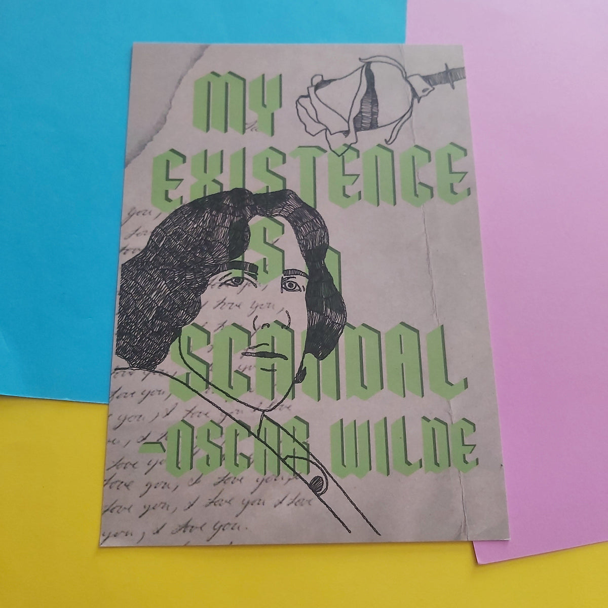 A print featuring a stylised portrait of Oscar Wilde with the text 'MY EXISTENCE IS A SCANDAL -OSCAR WILDE' in green, layered over sketchy black line art and cursive script on a beige background. The card is photographed on a bright yellow surface with blue and pink paper sheets partially visible behind it.