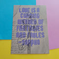 An art print featuring a quote by Sappho in lavender-purple text over a beige background with cursive script and a flower illustration, displayed on a colourful background of blue, pink, and yellow paper.
