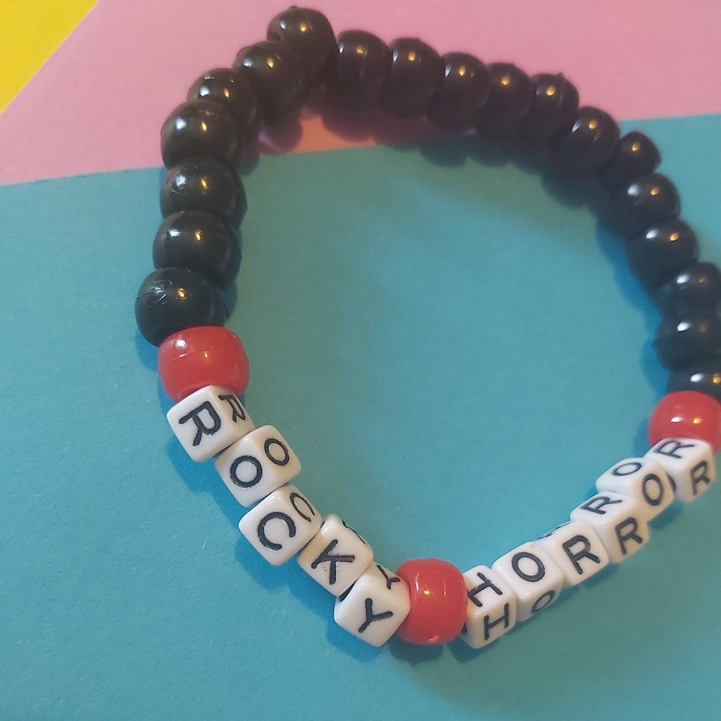 A beaded stretch bracelet with glossy black round beads, red beads, and white letter tiles spelling 'ROCKY HORROR' on a colourful blue, pink, and yellow background.