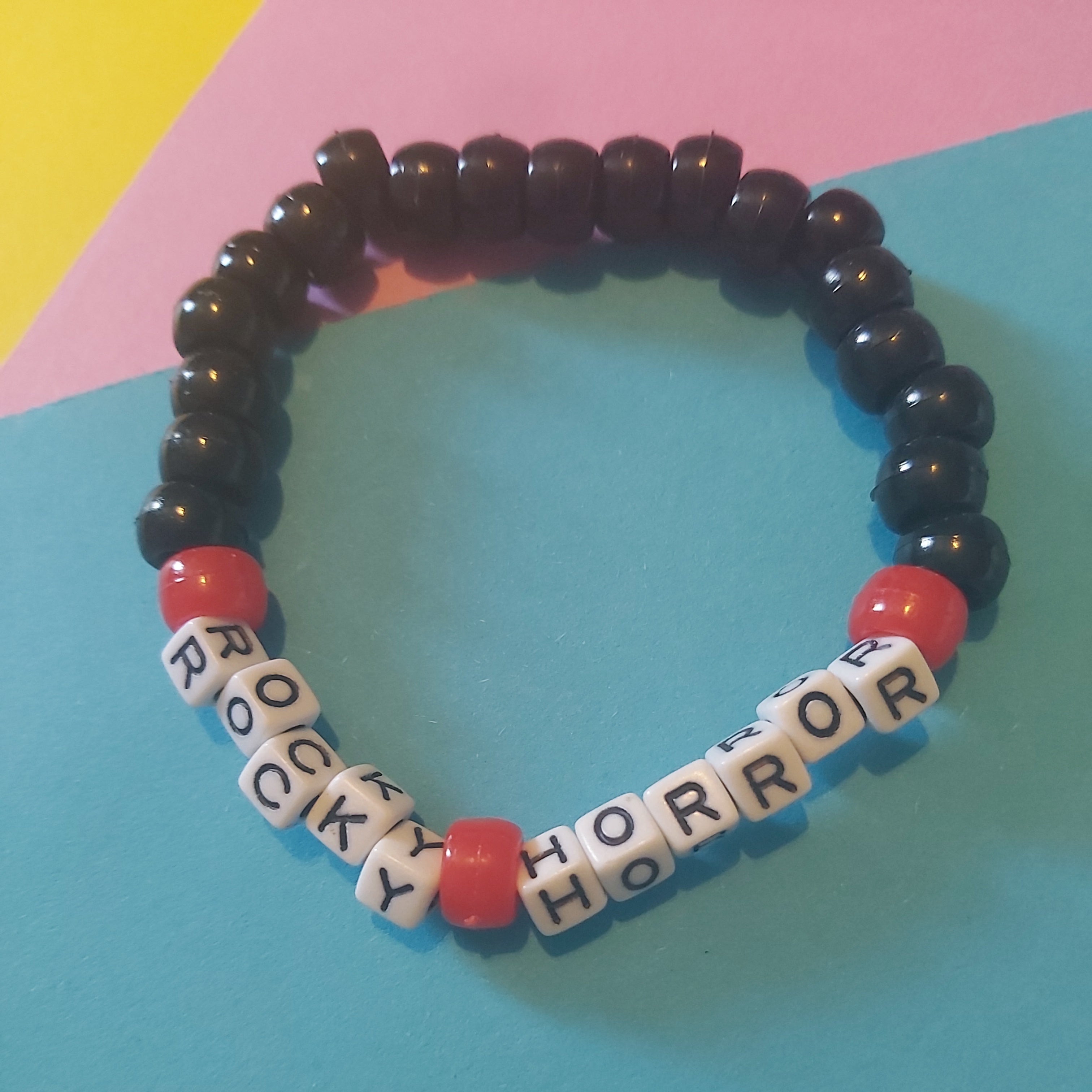 A beaded stretch bracelet featuring black round beads, red accent beads, and white letter tiles spelling 'ROCKY HORROR' on a colorful pastel background.