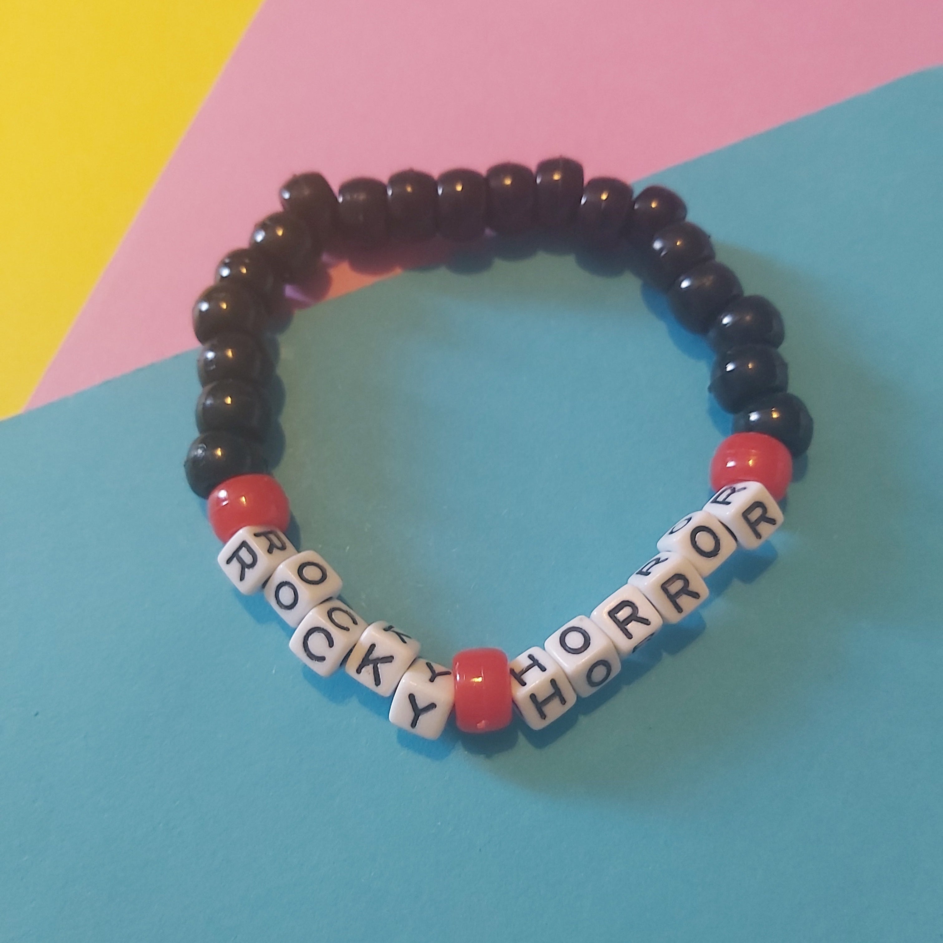 A beaded stretch bracelet featuring glossy black round beads, red cylindrical beads, and white square letter beads spelling out 'ROCKY HORROR'. The bracelet is displayed on a colorful, diagonally divided background of yellow, pink, and blue paper.